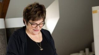 Eine Frau mit kurzen braunen Haaren und Brille, die ein schwarzes Oberteil und eine Halskette trägt, blickt nach unten, während sie im Haus steht.