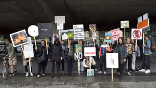Eine Gruppe von Menschen steht auf einem Bürgersteig und hält unter einem Betonvorsprung verschiedene Protestschilder mit unterschiedlichen Bildern, Symbolen und Botschaften hoch.
