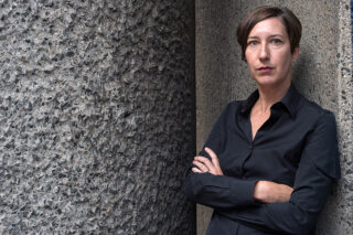 Eine Person mit kurzen braunen Haaren und einem schwarzen Hemd mit Knöpfen steht mit verschränkten Armen vor einer rauen Betonwand.