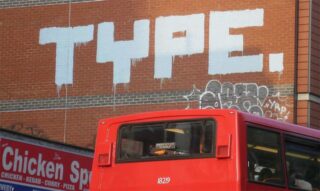 Auf eine Backsteinmauer über einem roten Bus und einem Ladenschild, das für Hühnchen, Kebab, Curry und Pizza wirbt, ist ein großes weißes Graffiti mit der Aufschrift „TYPE.“ gemalt.