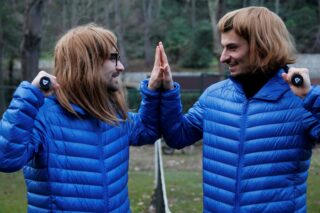 Zwei Menschen in blauen Pufferjacken und braunen Perücken geben sich im Freien ein High-Five, im Hintergrund sind Bäume und Gras zu sehen.