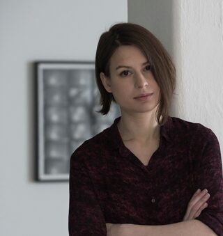 Eine Frau mit kurzen braunen Haaren und einem kastanienbraunen Hemd steht mit verschränkten Armen in einem Raum und lehnt an einer weißen Wand. Im Hintergrund hängt ein gerahmtes, verschwommenes Kunstwerk.