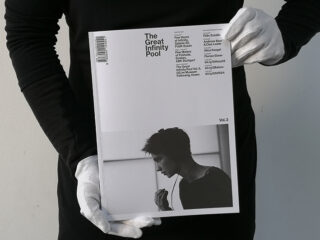 Eine Person mit weißen Handschuhen und schwarzer Kleidung hält eine Zeitschrift mit dem Titel „The Great Infinity Pool Vol. 3“, auf deren Cover ein Schwarzweißfoto einer Person im Profil zu sehen ist.