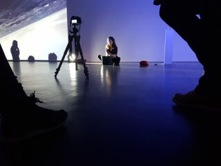 Eine Person sitzt in einem schwach beleuchteten Raum vor einer Projektion und einem Kamerastativ auf dem Boden. Im Vordergrund sind Silhouetten von Menschen und Schatten zu sehen.