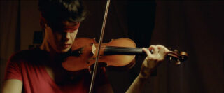 Eine Person mit roter Augenbinde und rotem Hemd spielt unter warmer Beleuchtung Geige.