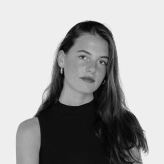 Eine junge Frau mit langem, glattem Haar und Creolen sowie einem ärmellosen schwarzen Top posiert vor einem schlichten, hellen Hintergrund.