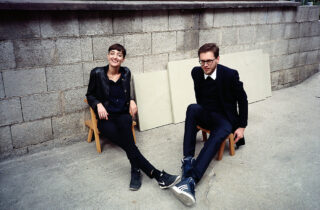 Zwei dunkel gekleidete Menschen sitzen im Freien auf kleinen Holzstühlen an einer Steinmauer, hinter ihnen lehnen helle Bretter.