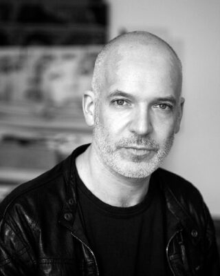 Ein Mann mit rasiertem Kopf und Bart trägt ein schwarzes Hemd und eine Lederjacke und blickt in einer sanft beleuchteten Innenumgebung in die Kamera.