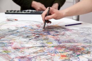 Eine Hand zeichnet mit einem Bleistift auf ein großes Blatt voller bunter Kritzeleien und Skizzen, während eine andere Person in der Nähe steht und im Hintergrund eine Tastatur bedient.