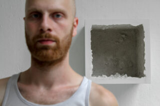 Ein Mann in einem weißen Tanktop steht vor einer weißen Wand, in die ein quadratischer, hohler Betonkasten eingelassen ist. Der Mann ist unscharf; der Kasten ist scharf.