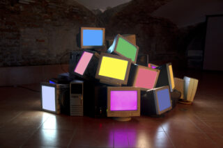 Ein Stapel alter Computermonitore und -türme auf einem gefliesten Boden, jeder Bildschirm zeigt eine andere Volltonfarbe an, vor dem Hintergrund einer Ziegelwand.