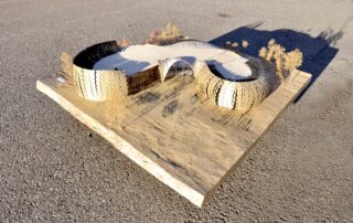 Auf einer flachen Außenfläche, umgeben von Miniaturbäumen, wird ein maßstabsgetreues Architekturmodell mit geschwungenen, strukturierten Wänden und offenem Mittelraum ausgestellt.