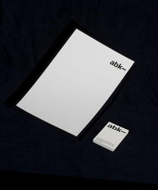 Auf einem dunklen Untergrund liegen ein leeres Blatt Papier und eine Visitenkarte, beide mit dem schwarzen „abk“-Logo versehen.