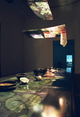 Ein langer Tisch mit Keramikschüsseln wird durch Deckenpaneele beleuchtet, die in einem schwach beleuchteten Raum bunte Lichtmuster auf die Oberfläche werfen.