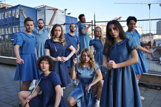 Eine Gruppe von zehn Menschen in blauen Kleidern posiert im Freien vor einer Baustelle, hält Golfschläger in den Händen und blickt ernst in die Kamera.