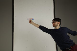 Eine Person mit Brille und dunklem Pullover wirft vor einer großen weißen Wand ein Papierflugzeug.
