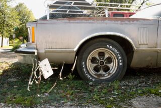 Ein rostiges Auto mit fehlendem Nummernschild an der Rückseite, das lose an Drähten in der Nähe des Hinterrads hängt, steht auf überwuchertem Kies.