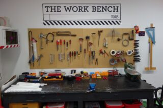 Eine Werkbank mit verschiedenen Handwerkzeugen, Klebeband und Zubehör, ordentlich auf einer Stecktafel angeordnet, unter einem Schild mit der Aufschrift „DIE WERKBANK-WERKZEUGE“.