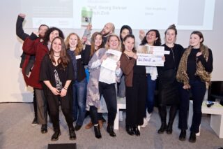 Eine Gruppe von Menschen posiert in einem Innenbereich, lächelt, hält Zertifikate und einen großen Scheck in den Händen und steht vor einem Bildschirm, auf dem Text angezeigt wird.