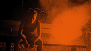 Eine Person sitzt nachts auf einer Bank neben einer leuchtend orangefarbenen Fackel, Rauch und Funken erhellen die Szene.