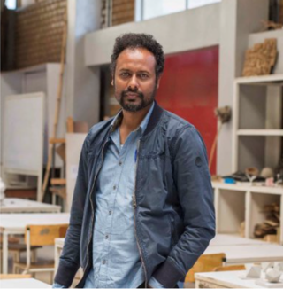 Ein Mann mit kurzen lockigen Haaren und Bart, der ein blaues Hemd und eine Jacke trägt, steht in einem hellen Raum mit Regalen, Tischen und Kunstbedarf im Hintergrund.