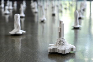 Eine Nahaufnahme einer weißen geometrischen Skulptur, die einer kleinen Stadtlandschaft ähnelt, mit ähnlichen abstrakten Teilen, die auf einer reflektierenden Oberfläche im Hintergrund verstreut sind.