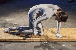 Eine Person in einem silbernen Ganzkörperanzug kniet auf einem Holzbrett und hält einen Hammer, auf dem Metallspäne verstreut liegen.