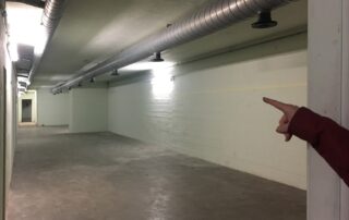 Die Hand einer Person zeigt auf einen leeren, gut beleuchteten Korridor mit Betonwänden, gefliestem Boden und Lüftungsschächten an der Decke.