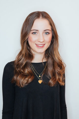 Eine junge Frau mit langem, braunem, welligem Haar trägt ein schwarzes Oberteil und zwei goldene Halsketten, eine davon mit einem Herzanhänger, und steht vor einem hellen Hintergrund.