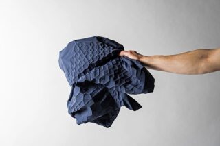 Eine Hand hält ein Stück dunkelblauen Stoff mit einem erhabenen geometrischen Sechseckmuster vor einem schlichten Hintergrund.