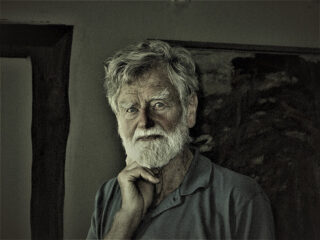 Ein älterer Mann mit grauem Haar und Bart steht im Haus. Er trägt ein Hemd mit Kragen und hat nachdenklich die Hand am Kinn.