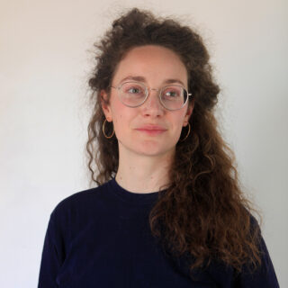 Eine Frau mit lockigem braunem Haar, Brille und Creolen steht vor einem schlichten hellen Hintergrund und trägt ein dunkles Hemd.