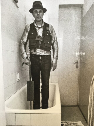 Ein Mann mit Hut, Weste und Jeans steht in einer Badewanne und hält einen stockähnlichen Gegenstand in einem gefliesten Badezimmer.