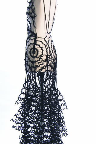 Ein transparentes Kleid mit schwarzen geometrischen und netzartigen Mustern, mit kreisförmigen Motiven und mehrlagigen Rüschen, von der Seite vor einem weißen Hintergrund fotografiert.