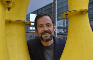 Ein Mann mit Bart und Schnurrbart lächelt, während er im Freien hinter einer großen gelben abstrakten Skulptur steht.