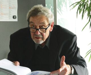 Ein älterer Mann mit Brille schaut beim Sprechen nach oben, gestikuliert mit einer Hand und hält mit der anderen ein offenes Buch. Er sitzt im Haus neben einer Pflanze und einem Wandkalender.