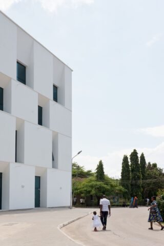 Ein Mann hält die Hand eines kleinen Kindes, während sie auf einem geschwungenen Gehweg neben einem modernen weißen Gebäude gehen, im Hintergrund sind Bäume und ein paar Menschen zu sehen.