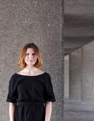 Eine Frau mit schulterlangem braunem Haar und einem schwarzen Kleid steht vor einer strukturierten Betonwand, im Hintergrund ist ein Korridor zu sehen.