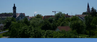 Eine kleine europäische Stadt mit Kirchtürmen, Gebäuden mit roten Dächern, dichten grünen Bäumen und einem Baukran unter einem klaren blauen Himmel.