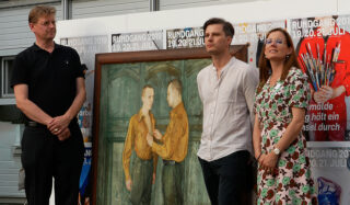 Drei Personen stehen neben einem gerahmten Gemälde, das zwei einander gegenüberstehende Männer zeigt; im Hintergrund sind Veranstaltungsplakate zum „Rundgang 2019“ zu sehen.