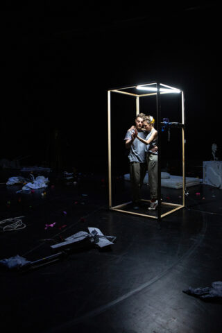 Zwei Personen stehen dicht beieinander in einem quadratischen, beleuchteten Rahmen auf einer dunklen Bühne, umgeben von verstreuten Requisiten und Trümmern.