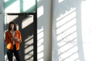 Eine Person in einer orangefarbenen Jacke steht mit einer Tasse in der Hand an einer Tür. Sonnenlicht wirft Schatten auf die Wand und im Hintergrund sind Skizzen zu sehen.