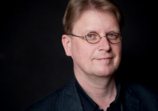 Ein Mann mit hellbraunem Haar, einer runden Brille und einem dunklen Blazer blickt vor einem schlichten dunklen Hintergrund in die Kamera.