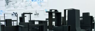 Auf einem Tisch sind mehrere abstrakte schwarze Architekturmodelle ausgestellt, im Hintergrund sind Skizzen und Zeichnungen an einer weißen Wand befestigt.