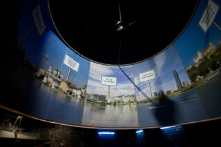 Auf einem gebogenen Panoramadisplay sind Stadtansichten mit beschrifteten Sehenswürdigkeiten und darüber ein Deckenventilator zu sehen, der von unten durch blaue Lichter beleuchtet wird.