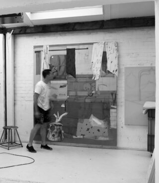 Eine Person steht vor einem großen, abstrakten Gemälde mit daran befestigten Stoffstücken, das in einem hellen, geräumigen Studio vor einer Ziegelwand ausgestellt ist.