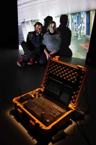 Zwei Personen sitzen an einer Wand, hinter ihnen wird ein Bild projiziert, während im Vordergrund ein Laptop in einem gelben Gehäuse eine Schnittstelle zur Gesichtserkennung anzeigt.