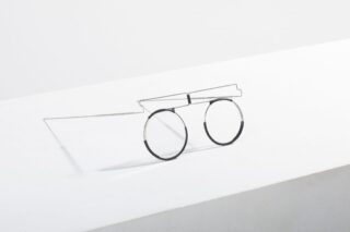 Eine minimalistische Brille mit Drahtgestell und runden Gläsern steht auf einer weißen Fläche vor einem schlichten weißen Hintergrund.