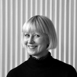 Eine Frau mit einem kurzen blonden Bob-Haarschnitt und einem schwarzen Rollkragenpullover steht lächelnd vor einem vertikal gestreiften Hintergrund.