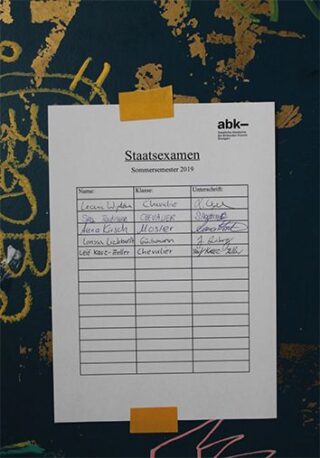 An einer Wand klebt ein Blatt mit der Aufschrift „Staatsexamen Sommersemester 2019“. Darin ist eine Tabelle mit Namen, Kursen und Unterschriften zu sehen. Vier Einträge sind handschriftlich eingetragen.